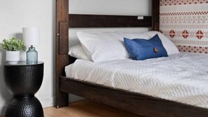 Una cama con ropa de cama blanca y una almohada azul, una mesita de noche con una planta y una lámpara, y una pared estampada al fondo.