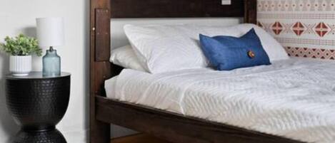 A bed with white bedding and a blue pillow, a small side table with a plant and a lamp, and a patterned wall in the background.