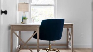 Una habitación con un escritorio de madera, una silla azul, una lámpara y un ventanal con vistas a árboles.