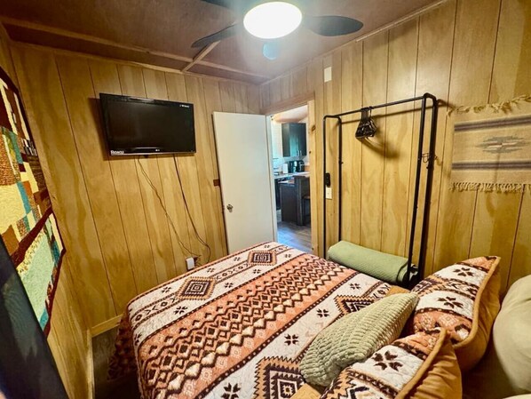 Une pièce chaleureuse aux murs en bois, avec un lit recouvert d’une literie à motifs, un ventilateur de plafond et un téléviseur fixé au mur.