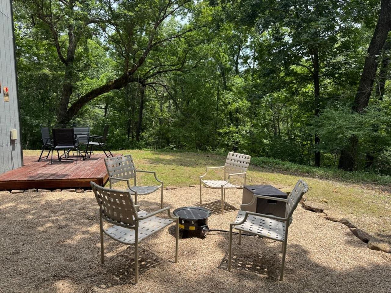 An outdoor seating area with metal chairs and a table, surrounded by trees.