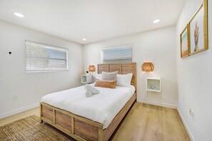 Een slaapkamer met een houten bed, witte beddengoed, twee nachtkastjes met lampen, en een groot raam met zonwering.