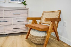 Een houten stoel met een geweven rugleuning en zitvlak, geplaatst op een geweven poef, naast een licht houten dressoir met lades.