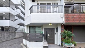 Een meerlaags woongebouw met een balkon, een voordeur en een klein buiten zitgedeelte met een bank en potplanten.