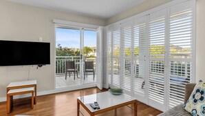 A living room with a flat-screen TV, a coffee table, and a sofa. There is a balcony with white shutters and a view of greenery. - The Pool Resort Kingscliff - One Bedroom (7106) - Holiday Management Co. (Tweed)