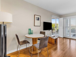 A modern living room with a round wooden table, two chairs, a TV mounted on the wall, and a balcony with sliding doors.