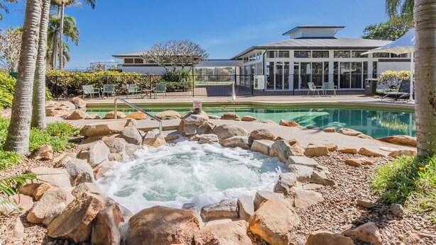 A pool area with a hot tub surrounded by rocks, a building with large windows, and palm trees. - The Pool Resort Kingscliff - One Bedroom (7106) - Holiday Management Co. (Tweed)