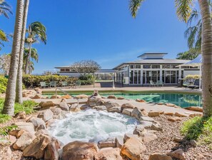 A pool area with a hot tub surrounded by rocks, a building with large windows, and palm trees.