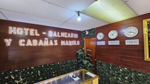 Vestíbulo de hotel con una pared de madera que muestra el nombre 'HOTEL - BALNEARIO Y CABANAS MANOLO'. Hay relojes mostrando diferentes horas y un letrero que indica 'TOKIO'.