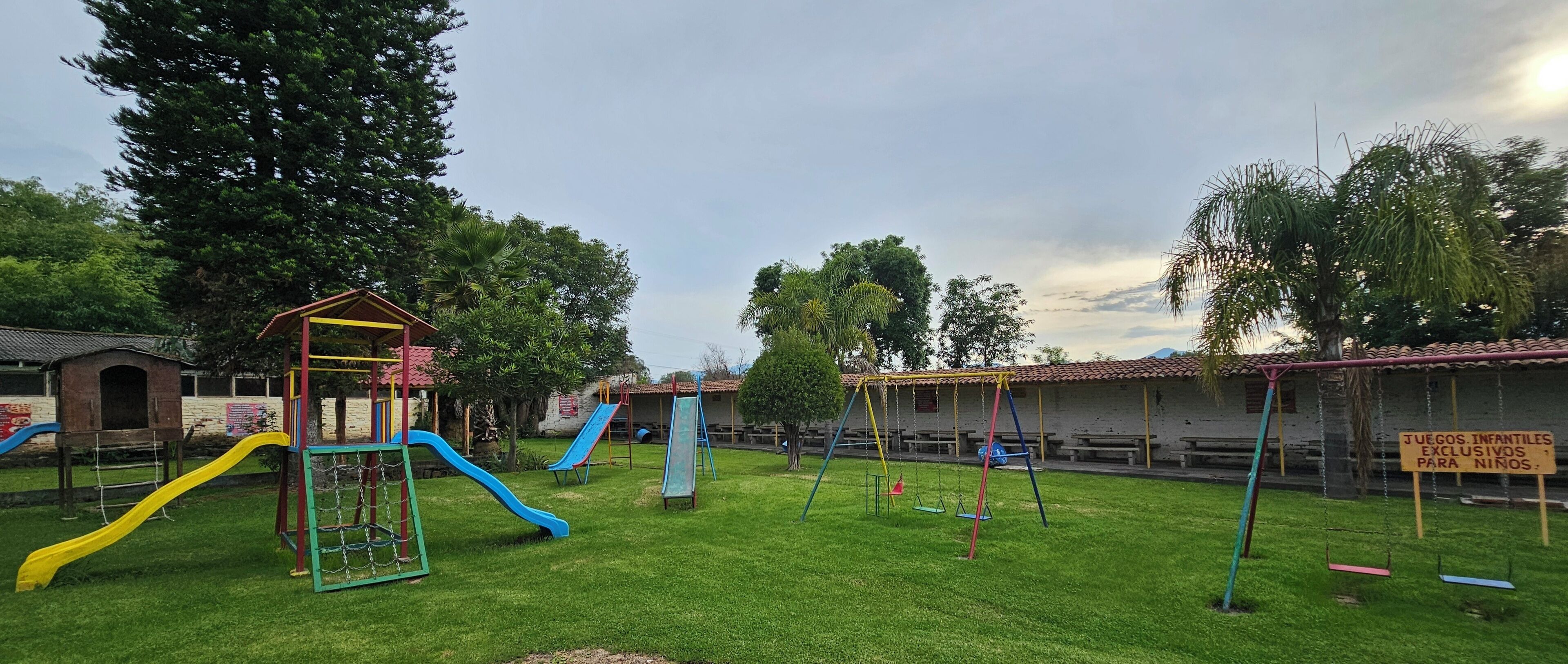 A playground with slides and swings, surrounded by trees and a building with a sign.