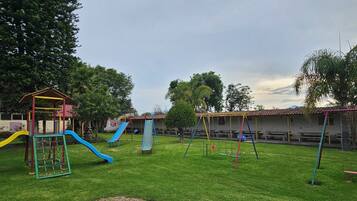 A playground with slides and swings, surrounded by trees and a building with a sign.