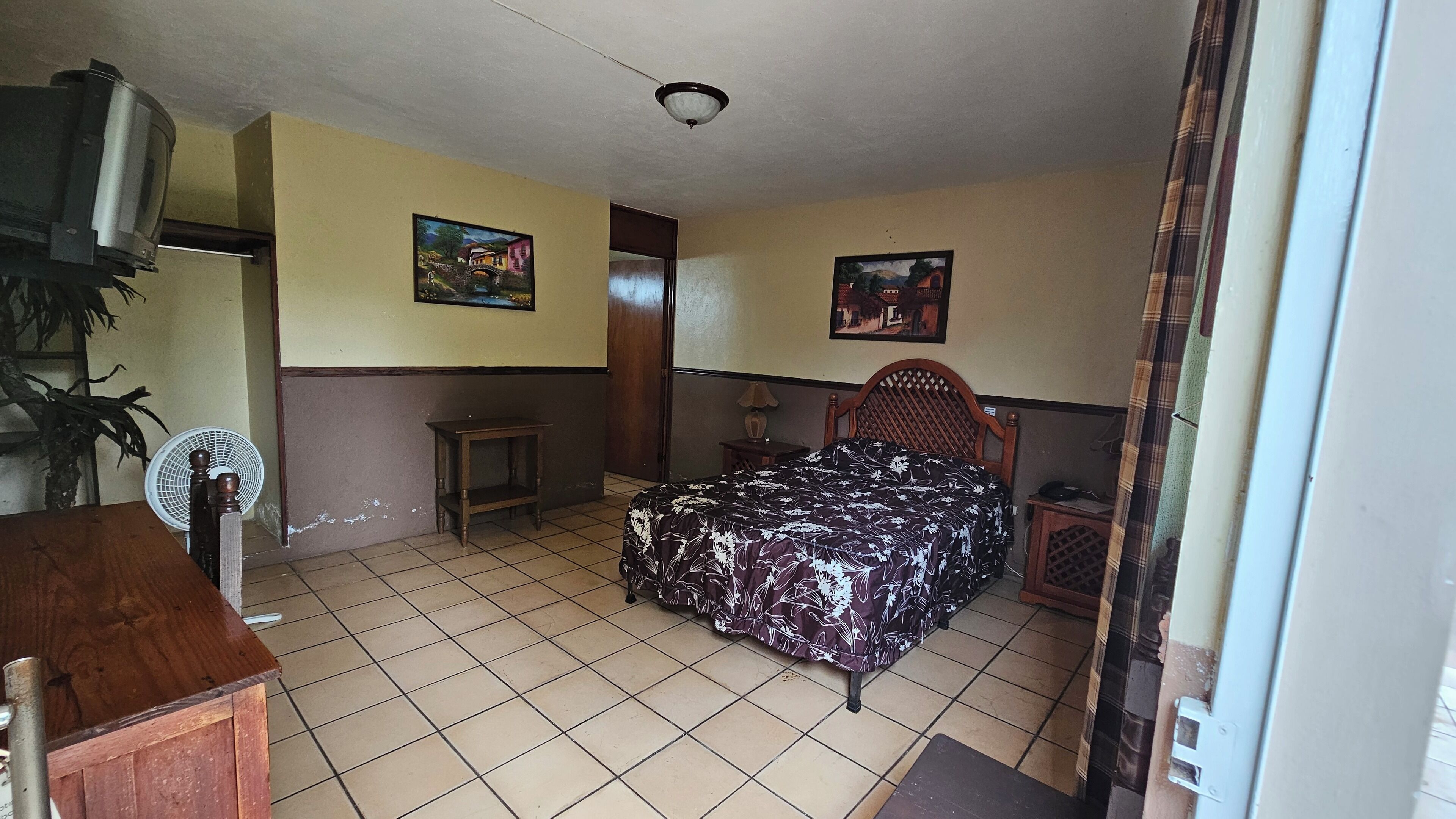 A small, simple room with a bed, a fan, a small table, and a TV.