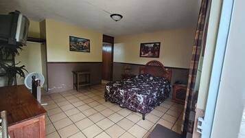 A small, simple room with a bed, a fan, a small table, and a TV.