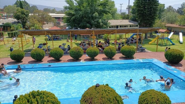 Una piscina con gente nadando y jugando, rodeada por una zona de estar cubierta y pequeños árboles.