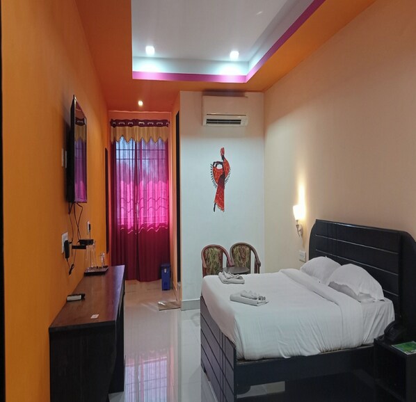 A hotel room with a bed, bedside table, and a desk with a chair. There is a window with curtains, a wall-mounted TV, and a wall decoration. - Joe Beach Resort (Tirupporur)