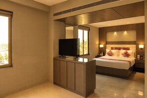 Una habitación de hotel moderna con un televisor de pantalla plana sobre una consola de madera, una cama grande con almohadas estampadas y una mesita de noche con lámpara.