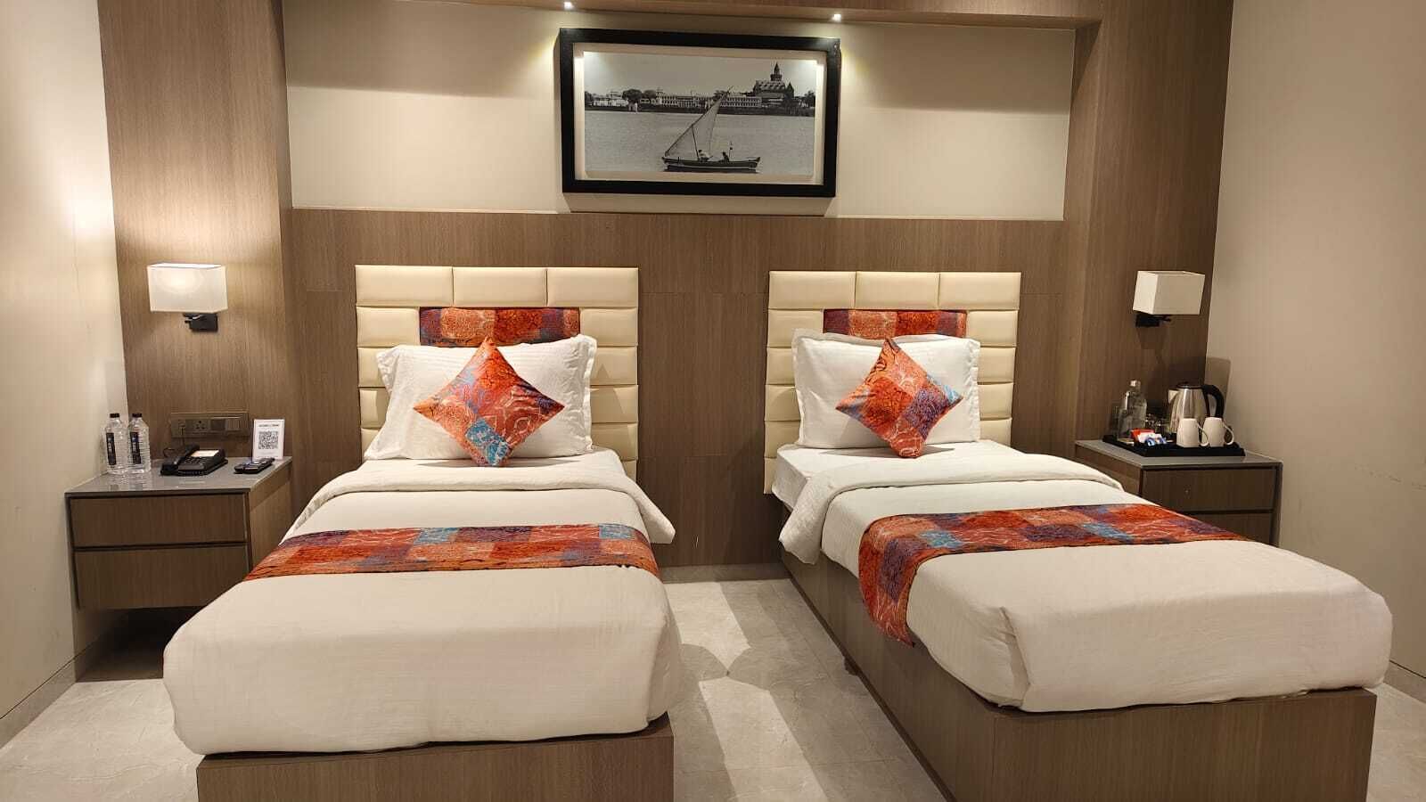 A hotel room with two beds, a framed picture above, bedside tables with lamps, and a small table with a kettle and cups.