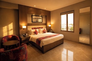 A hotel room with a large bed, two bedside tables, a chair, a window with a city view, and a framed picture on the wall. - LA CASA DE RANN (Bhuj)