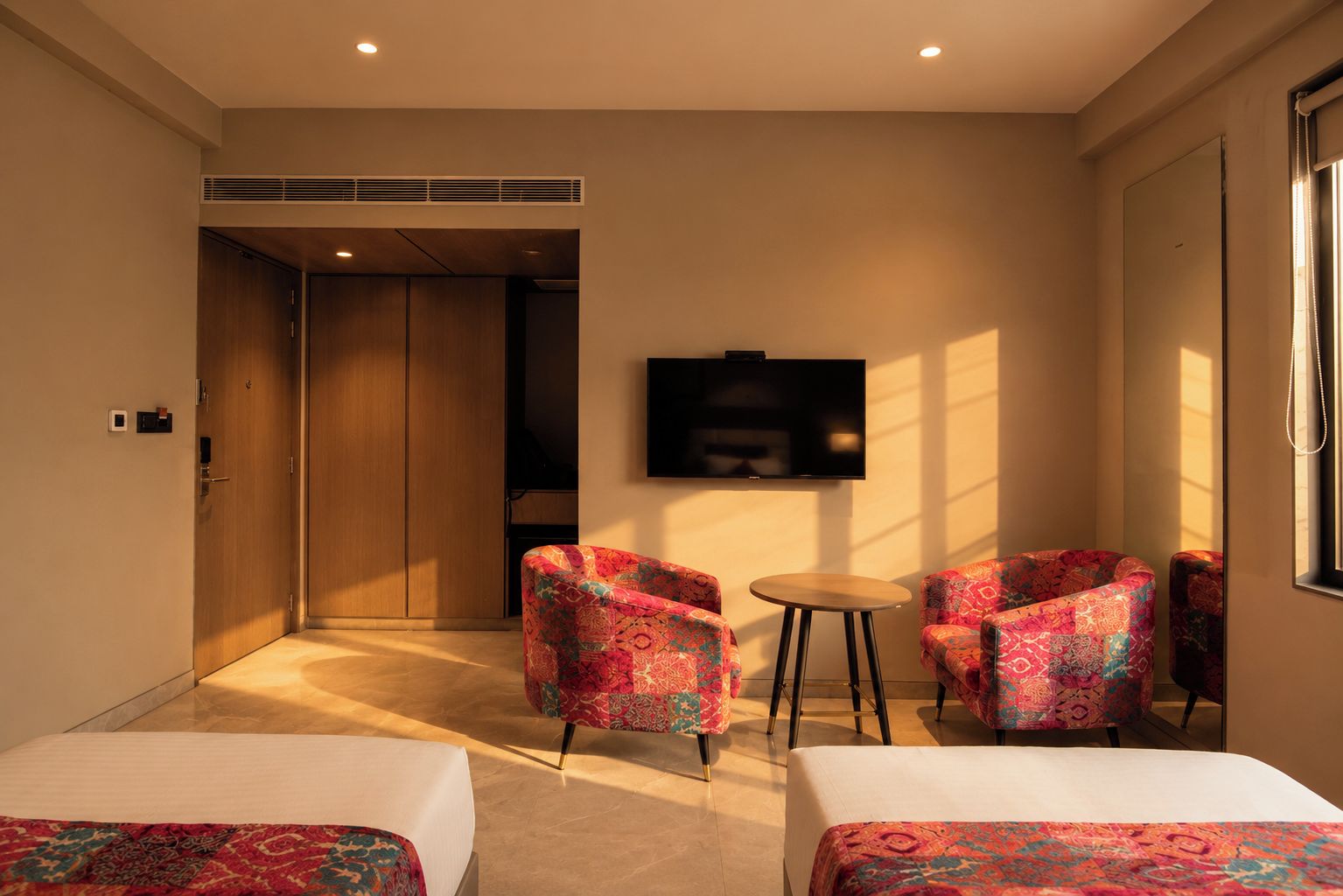 A hotel room with two beds, a TV, a small table, and two patterned armchairs.