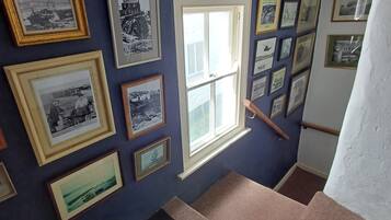Ein Zimmer mit einer dunkelblauen Wand, die mit zahlreichen gerahmten Fotografien geschmückt ist. Ein Fenster mit Holzrahmen ist sichtbar, und eine Holzhandrail verläuft entlang der Wand.