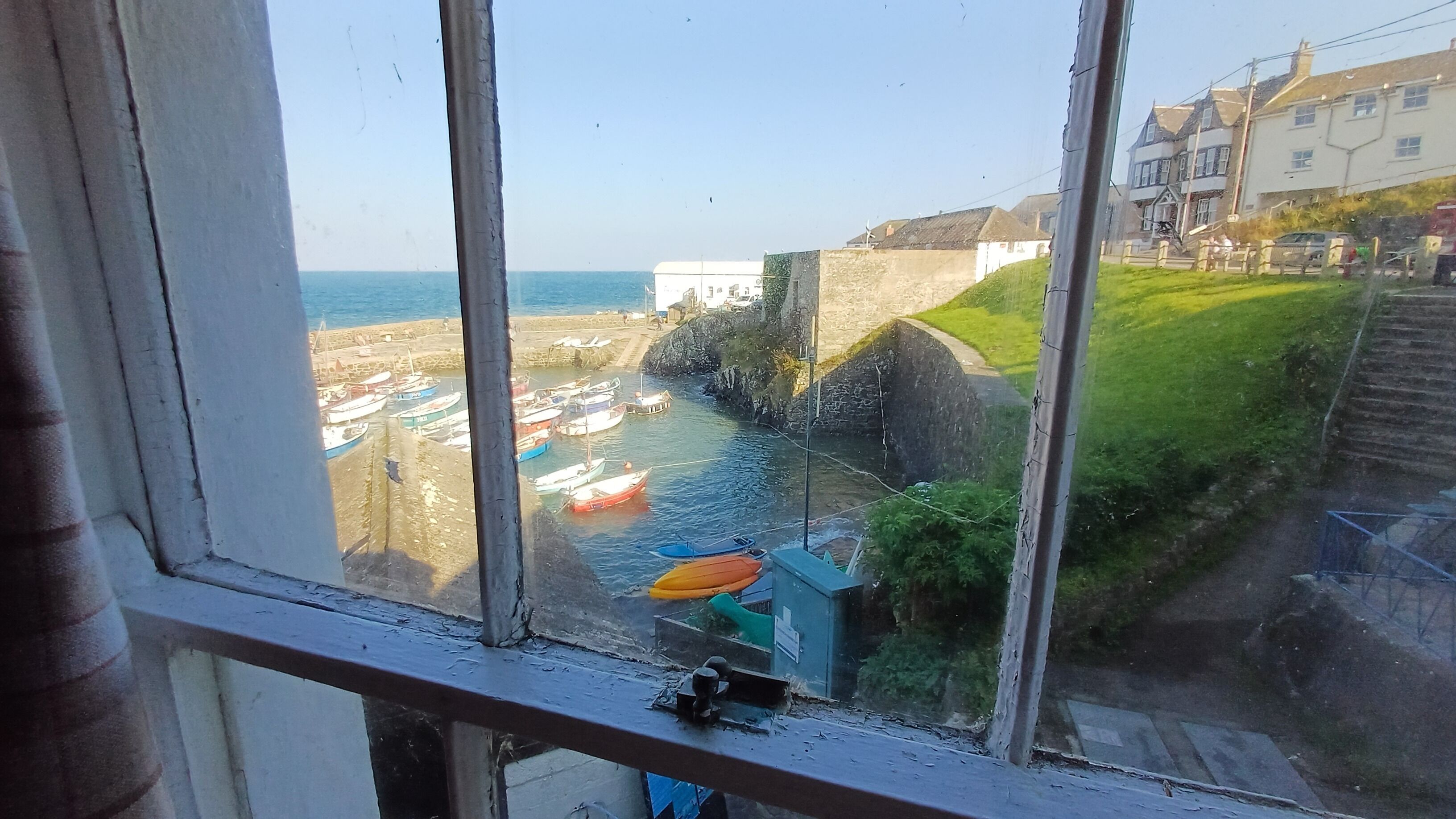 Blick aufs Meer vom Fenster aus, mit Blick auf einen Hafen, Schiffe, eine Steinmauer und Gebäude.