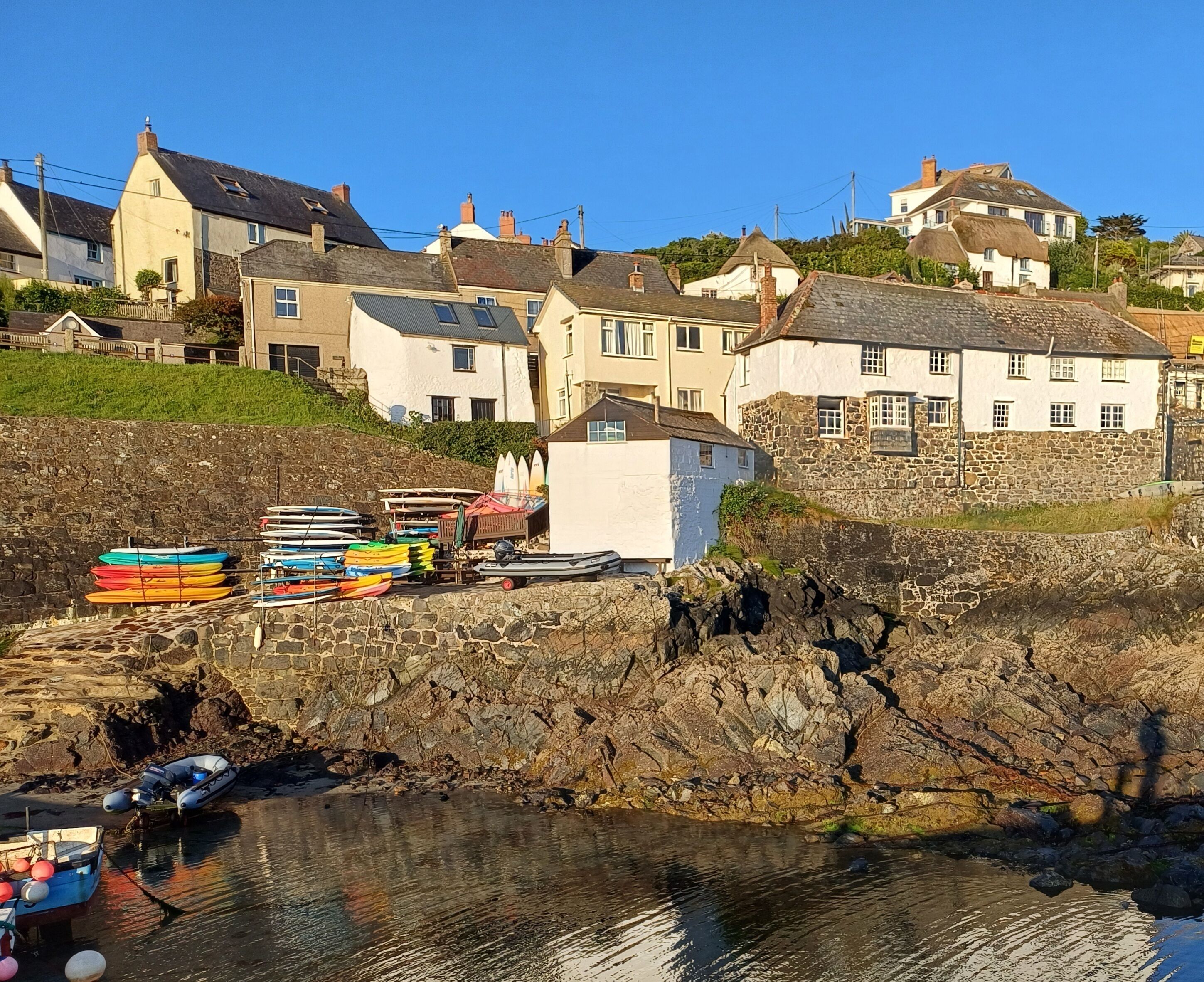 A coastal town with stone buildings, a rocky shoreline, and a row of colorful kayaks and a small boat docked at the water's edge.