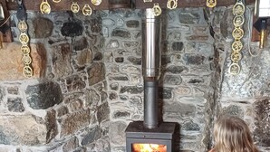 A stone fireplace with a modern wood stove, a mantel with decorative items, and a person sitting in front of it. - The Old Post Office (Coverack)