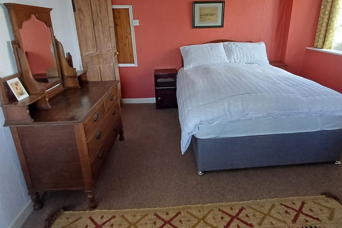 A bedroom with a wooden dresser, a bed with white bedding, a red wall, and a framed picture above the bed.