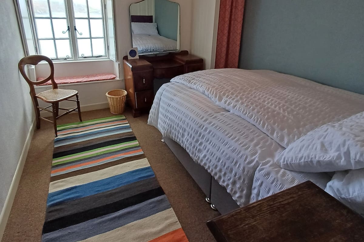 A bedroom with a bed, a chair, a small table, a mirror, a window, and a striped rug.