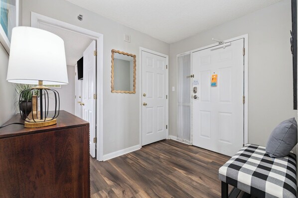 A hallway with wooden flooring, white doors, a mirror, a lamp, and a checkered bench.