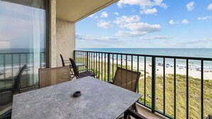 A balcony with a table and chairs overlooking a beach with umbrellas and people. - Oceanfront Luxury 4BR | Sleeps 10 | Beach Club II (North Myrtle Beach)