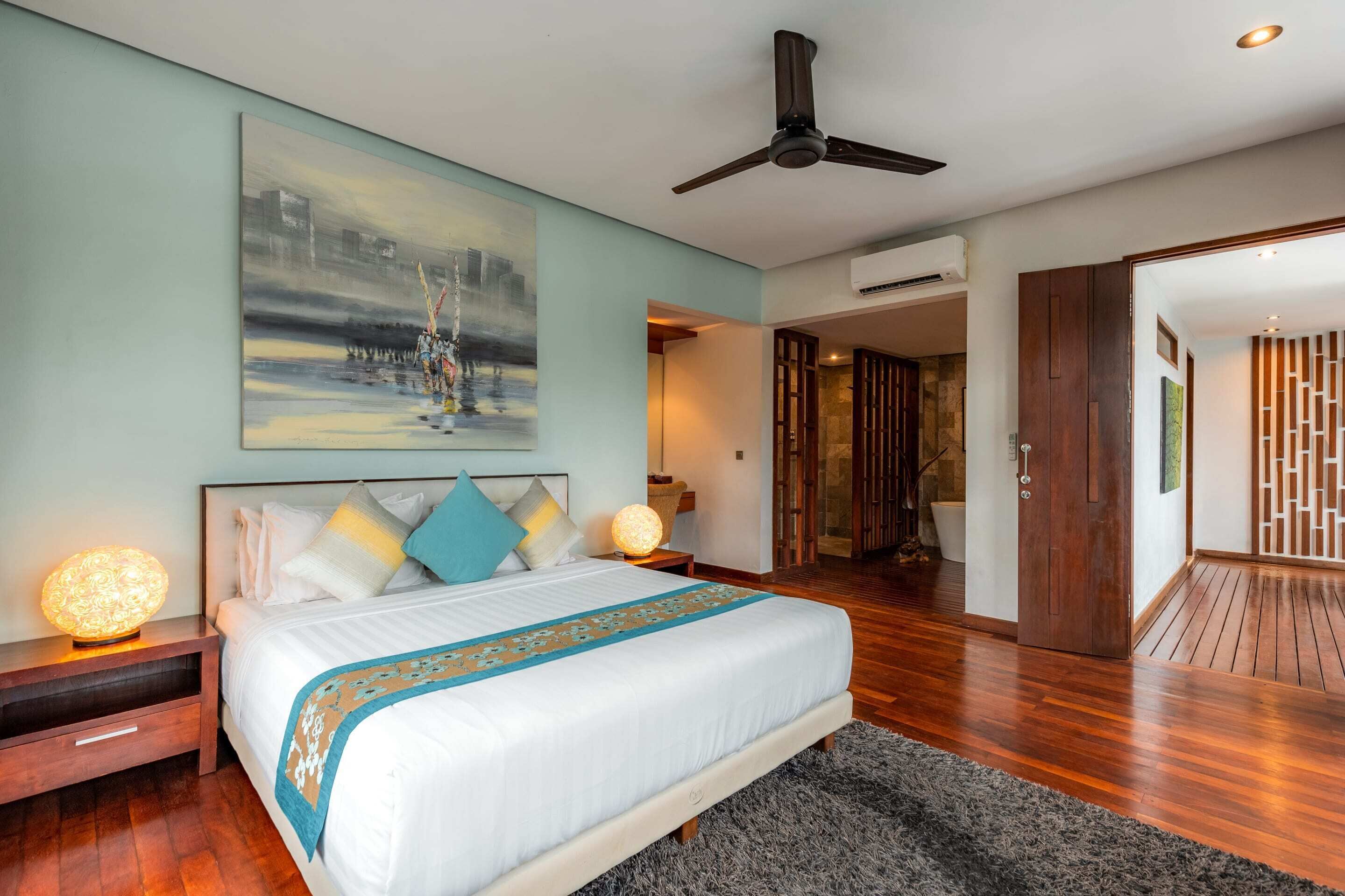 A modern hotel room with a large bed, a ceiling fan, and a painting on the wall.
