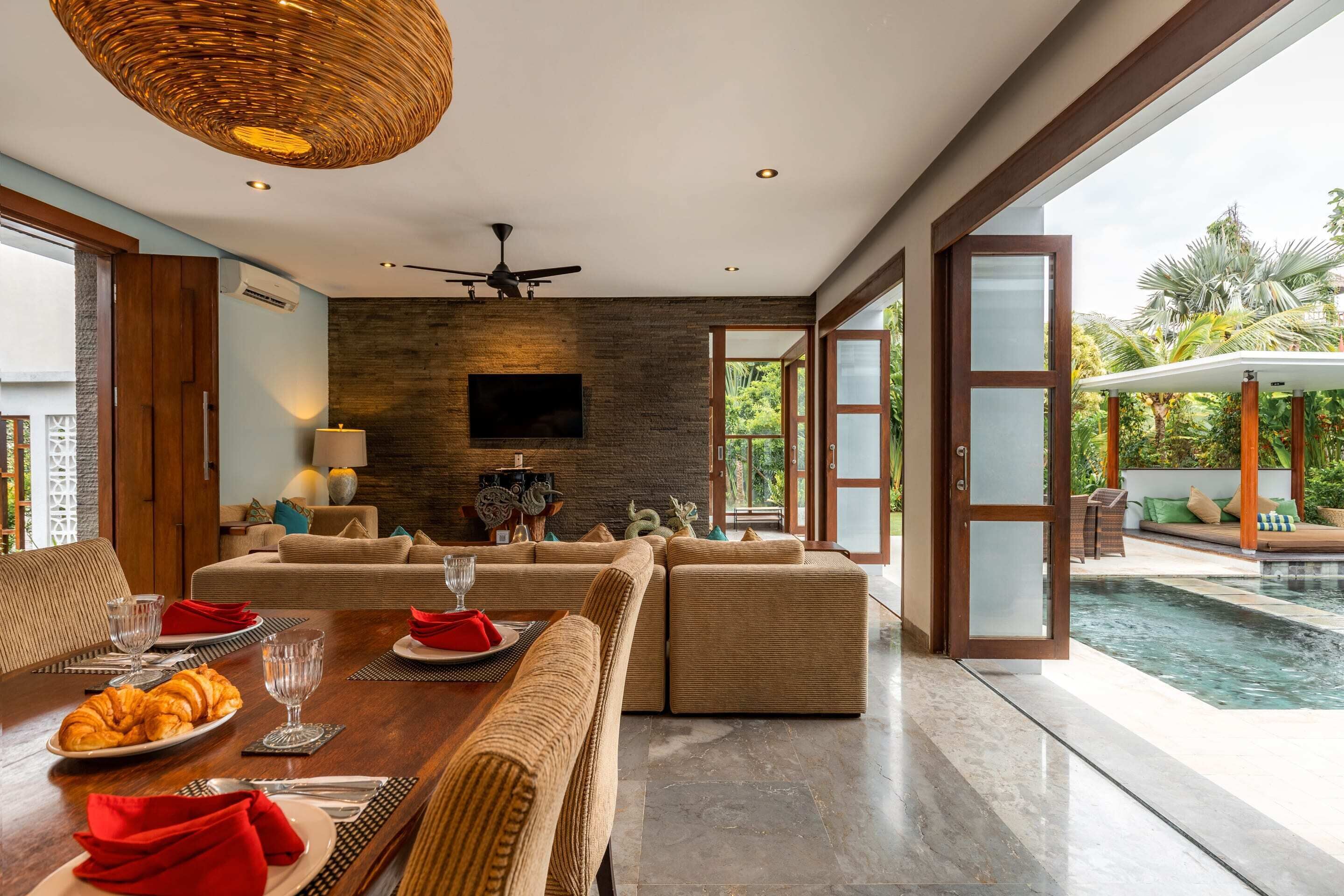 A modern living room with a large dining table, comfortable seating, a flat-screen TV, and a view of a swimming pool.