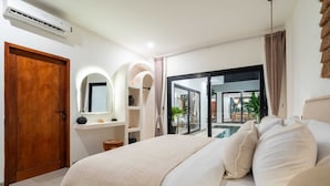 A modern bedroom with a large bed, a wooden door, a built-in shelf with decorative items, and a view of a pool and outdoor area. - Villa Lu'um (Canggu)