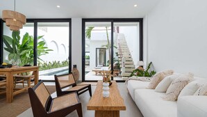 A modern living room with a wooden coffee table, white sofa, and wicker chairs. A staircase leads to an outdoor area with plants and a pool. - Villa Lu'um (Canggu)