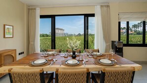 A dining area with a large table set for four, large windows with curtains, and a view of a field and building. - Villa Leblanc (Canggu)