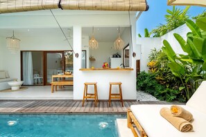 A modern outdoor pool area with a wooden deck, a bar with stools, and a seating area with a sofa and a table. - Villa Isla (Tabanan)