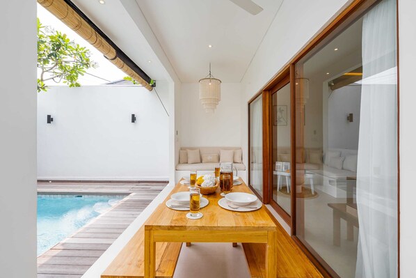 A dining area with a wooden table set for four, a pool in the background, and a modern interior with a sofa and a large window. - Villa Isla (Tabanan)