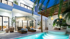 A modern outdoor pool area with lounge chairs and umbrellas, surrounded by large potted plants and a stone wall. - Villa Irie (Cemagi)