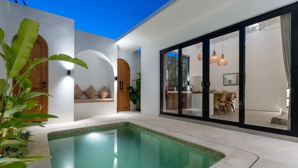 A modern indoor pool area with a clear pool, wooden doors, and a dining area with natural light. - Villa Delia (Munggu)