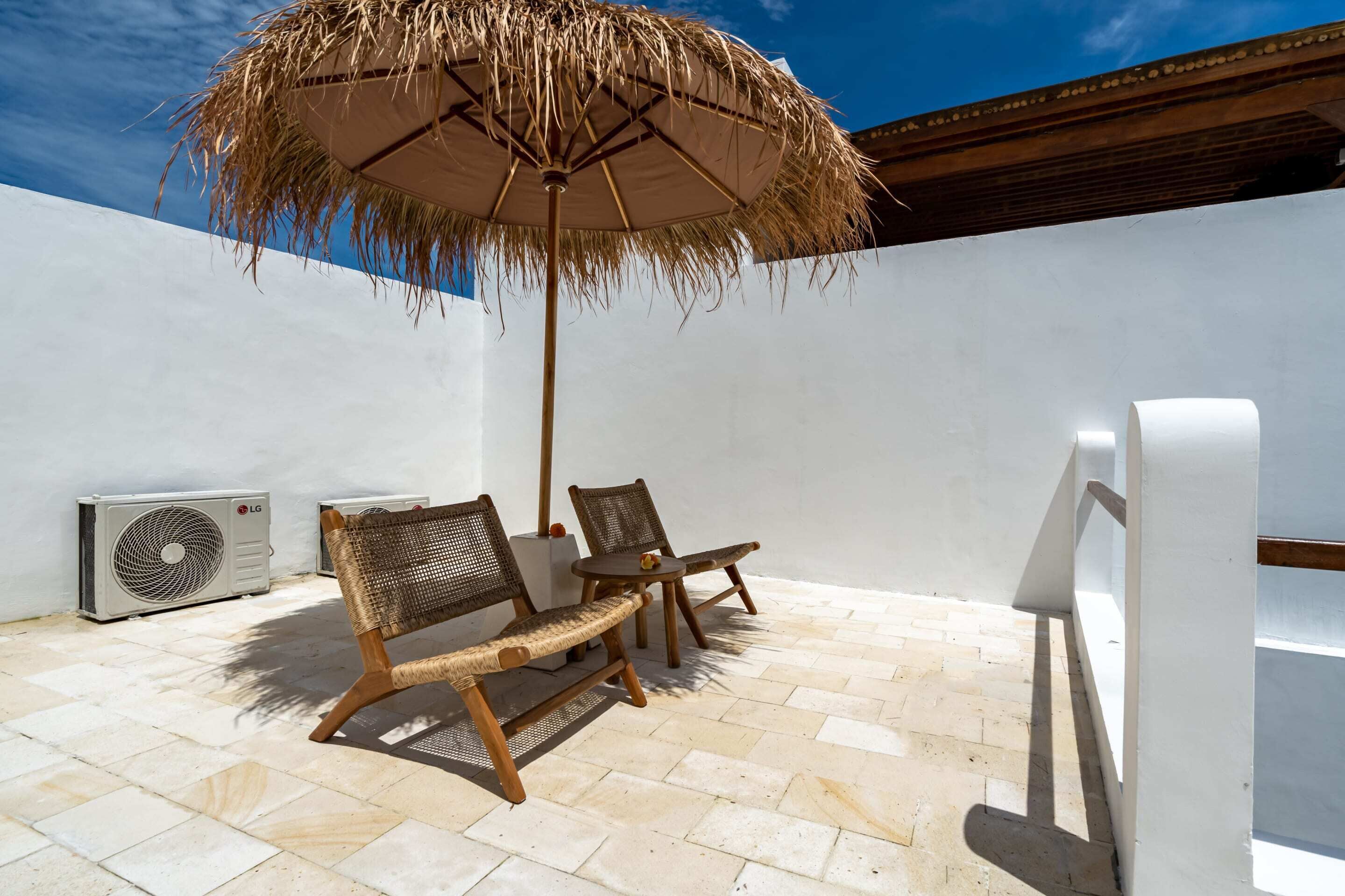 A patio with two wicker chairs, a straw umbrella, and an air conditioning unit.
