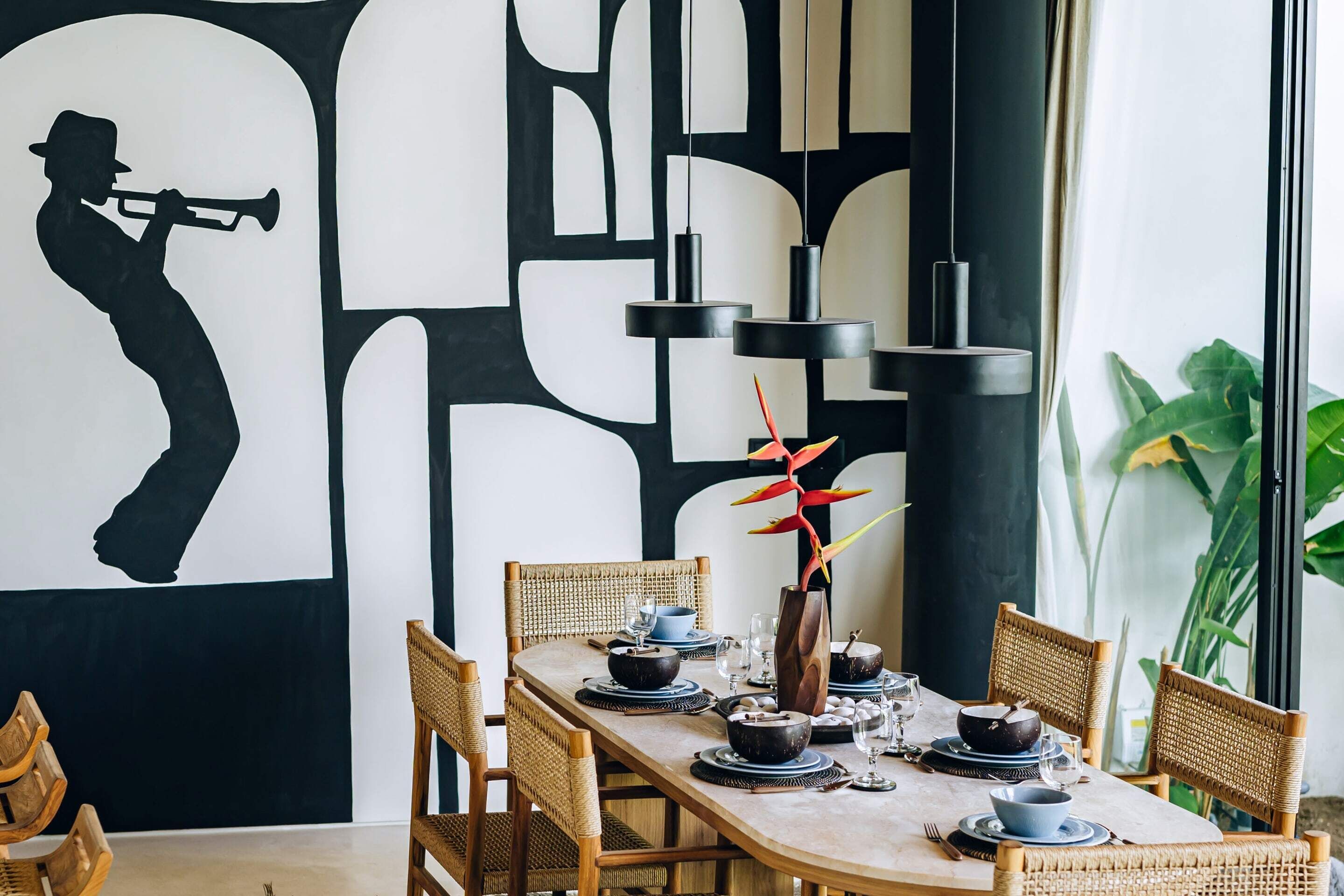 A dining area with a long table set for four, surrounded by wicker chairs. A large abstract painting is on the wall behind the table.