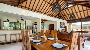 Un comedor espacioso con techo alto y vigas de madera, que presenta una larga mesa de madera dispuesta para una comida, rodeada de sillas de madera.