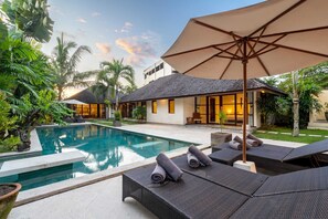 A poolside area with lounge chairs and an umbrella, surrounded by lush greenery and a building with large windows. - Villa Alroda Antik (Canggu)