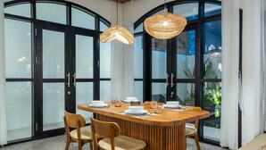 A dining area with a wooden table set for four, surrounded by chairs, with large windows and black-framed glass doors. - Villa Ambrogio A (Canggu)