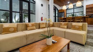 En moderne hotellobby med en stor beige hjørnesofa, et træsofabord med en potteplante, og et køkkenområde i baggrunden.