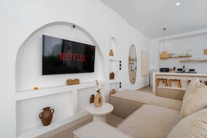 Una sala de estar moderna con un televisor de pantalla plana grande montado en una pared blanca, un sofá, una mesita redonda con un jarrón y una zona de cocina al fondo.