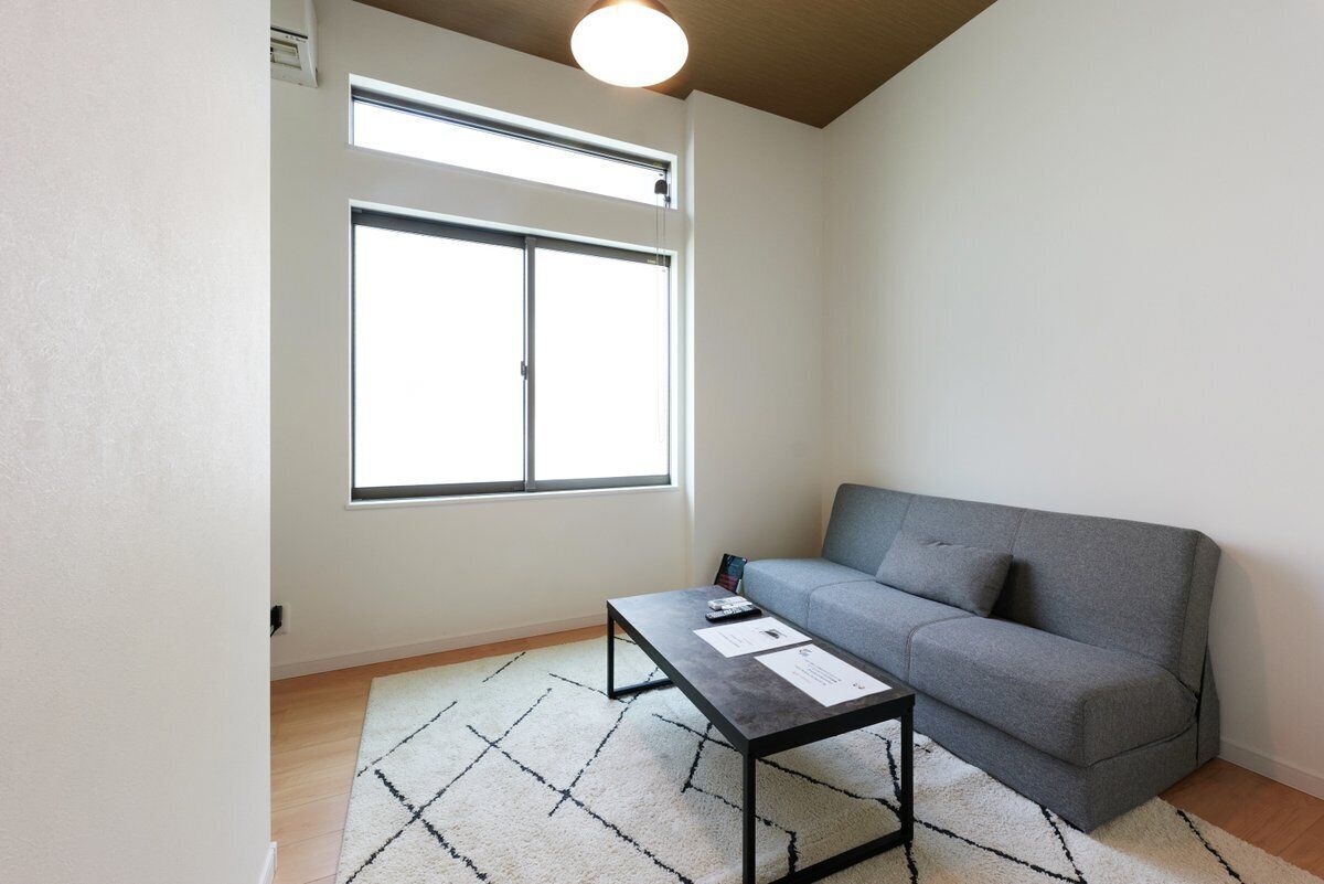 Ein Wohnzimmer mit grauem Sofa, einem schwarzen Couchtisch und einem gemusterten Teppich. Ein großes Fenster sorgt für natürliches Licht.