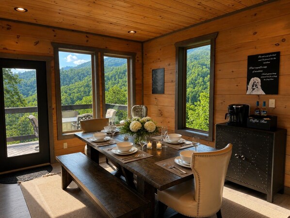 A wooden interior with a dining table set for four, a coffee bar, and a view of a forested mountain.