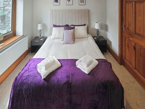 Habitación de hotel con una cama grande, dos mesitas de noche, un ventanal con vistas a muros de piedra y una puerta de madera.